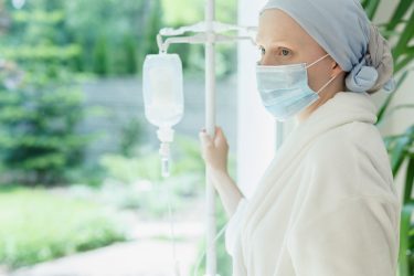 Woman wearing a head scarf connected to an IV drip looking out a window - idea of locked-down/shielding cancer patients