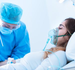 Young woman wearing an oxygen mask on a hospital bed, with doctor in blue PPE checking her IV - COVID-19 ARDS patient