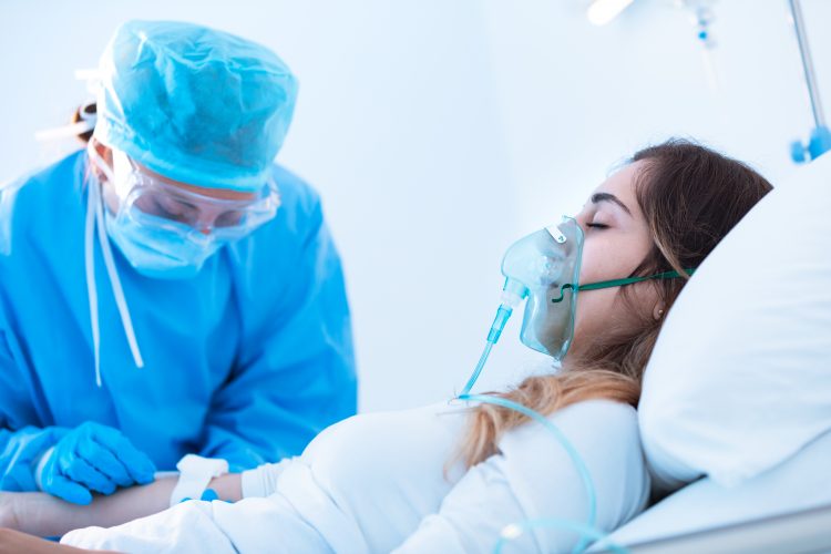 Young woman wearing an oxygen mask on a hospital bed, with doctor in blue PPE checking her IV - COVID-19 ARDS patient