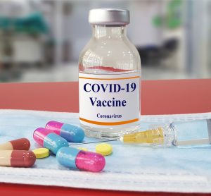 Vial labelled 'COVID-19 vaccine' surrounded by different coloured capsules and pillls and a syringe