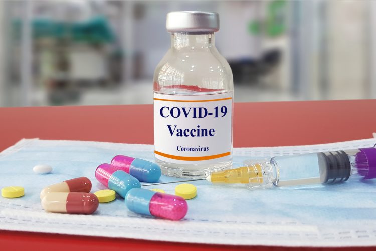Vial labelled 'COVID-19 vaccine' surrounded by different coloured capsules and pillls and a syringe