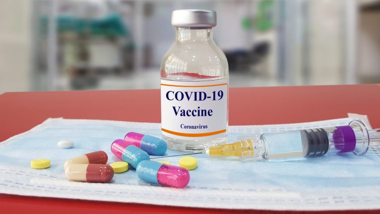 Vial labelled 'COVID-19 vaccine' surrounded by different coloured capsules and pillls and a syringe