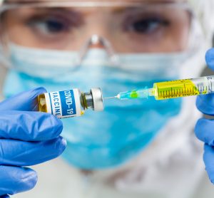 Doctor in full PPE drawing yellow liquid into a syringe from a vial labelled 'COVID-19 Vaccine'