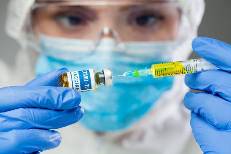 Doctor in full PPE drawing yellow liquid into a syringe from a vial labelled 'COVID-19 Vaccine'