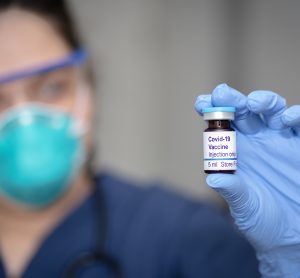 doctor in a mask holding a vial labelled 'COVID-19 Vaccine'