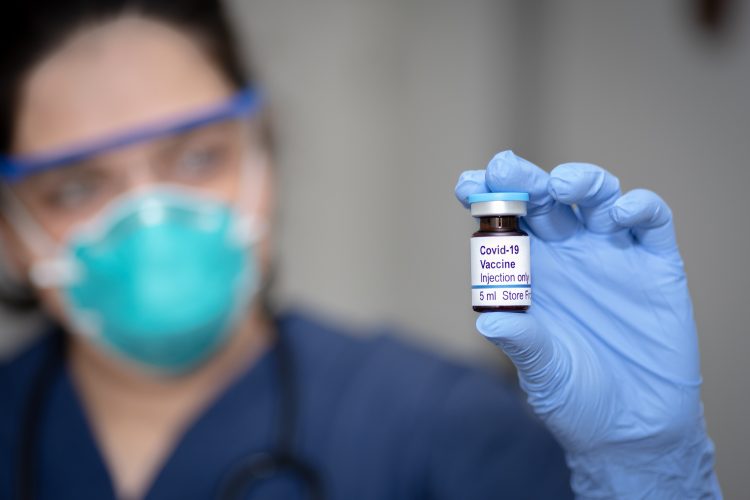 doctor in a mask holding a vial labelled 'COVID-19 Vaccine'