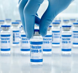 Hand of male doctor wearing medial glove holding covid 19 corona virus vaccine vial bottle for injection labelled 'COVID-19 vaccine' with a line up of similar vials in the background