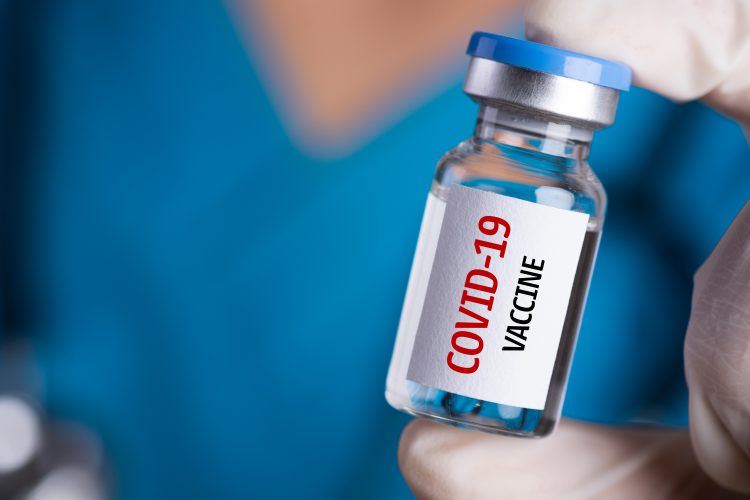 gloved hand holding a vial labelled 'COVID-19 vaccine'