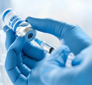 Close up of the gloved hands of a doctor drawing a vaccine dose into a syringe from a vial labelled 'COVID-19 VACCINE'