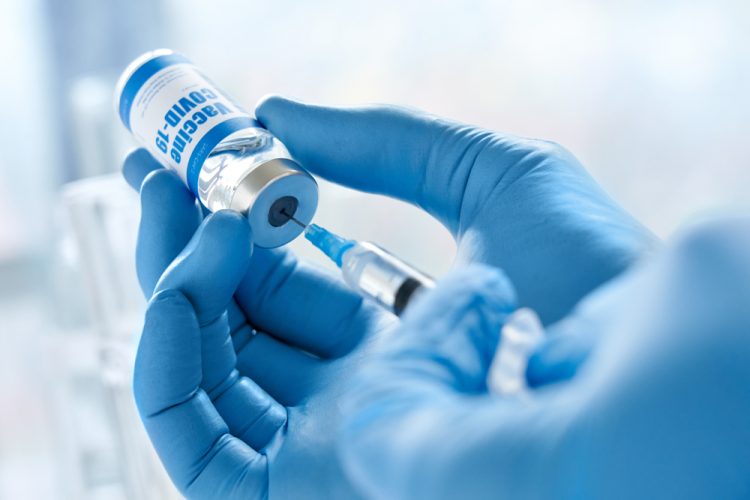 Close up of the gloved hands of a doctor drawing a vaccine dose into a syringe from a vial labelled 'COVID-19 VACCINE'