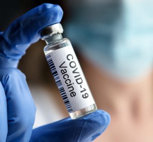 Doctor's gloved hand holding up a glass vial labelled 'COVID-19 Vaccine' with background blurred