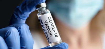 Doctor's gloved hand holding up a glass vial labelled 'COVID-19 Vaccine' with background blurred