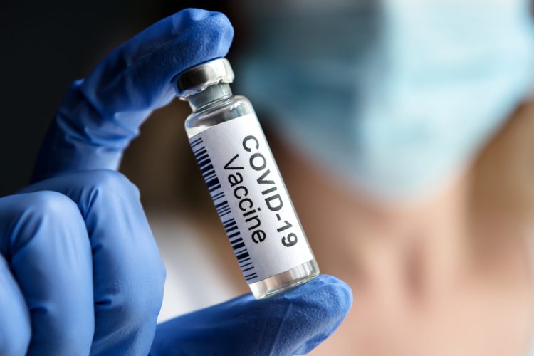 Doctor's gloved hand holding up a glass vial labelled 'COVID-19 Vaccine' with background blurred