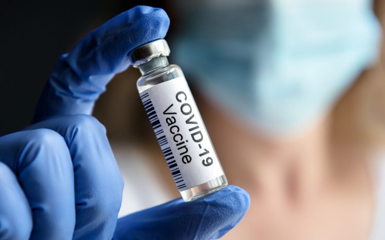 Doctor's gloved hand holding up a glass vial labelled 'COVID-19 Vaccine' with background blurred