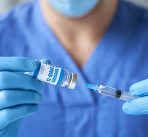 doctor wearing blue uniform, mask, medical gloves holding syringe drawing liquid into a syringe from a vial labelled 'COVID-19 coronavirus vaccine' preparing for injection.