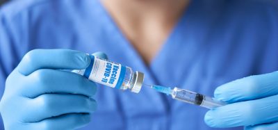 doctor wearing blue uniform, mask, medical gloves holding syringe drawing liquid into a syringe from a vial labelled 'COVID-19 coronavirus vaccine' preparing for injection.