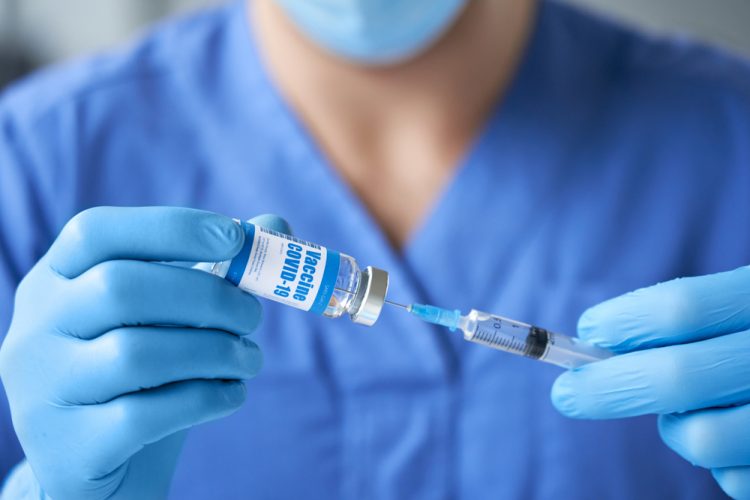 doctor wearing blue uniform, mask, medical gloves holding syringe drawing liquid into a syringe from a vial labelled 'COVID-19 coronavirus vaccine' preparing for injection.