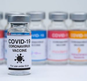 Covid 19 vaccine bottles in the background with the logos of different pharmaceutical companies and in focus a vaccine bottle labelled 'COVID-19 Vaccine' no company logo