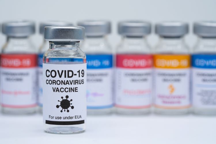 Covid 19 vaccine bottles in the background with the logos of different pharmaceutical companies and in focus a vaccine bottle labelled 'COVID-19 Vaccine' no company logo