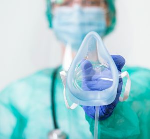 Female doctor in full personal protective equipment holding an oxygen mask towards the camera - idea of respiratory failure due to COVID-19/inhaled treatment for COVID-19