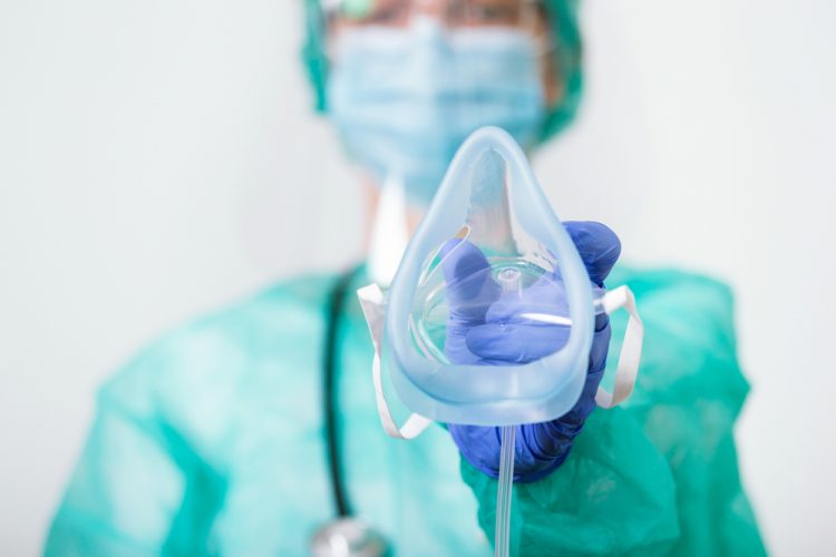 Female doctor in full personal protective equipment holding an oxygen mask towards the camera - idea of respiratory failure due to COVID-19/inhaled treatment for COVID-19