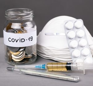 a pile of face masks next to a syringe, a thermometer and a jar of coins labelled 'COVID-19'