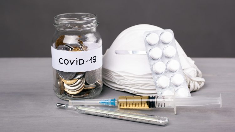 a pile of face masks next to a syringe, a thermometer and a jar of coins labelled 'COVID-19'