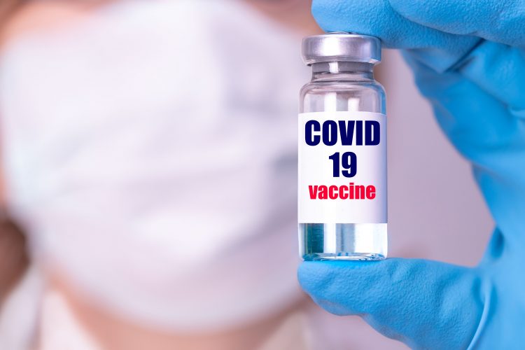 doctor wearing face mask holding a vial labelled 'COVID-19 Vaccine'