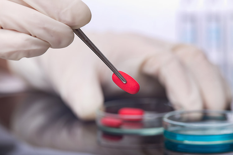 Close up of scientist's hands holding red pill with tweezers above petri dish.