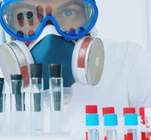 Scientist in respirator, goggles and other protective gear looking at camera over a lab bench of sample tubes - idea of HPAPIs