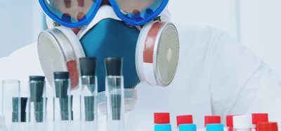 Scientist in respirator, goggles and other protective gear looking at camera over a lab bench of sample tubes - idea of HPAPIs