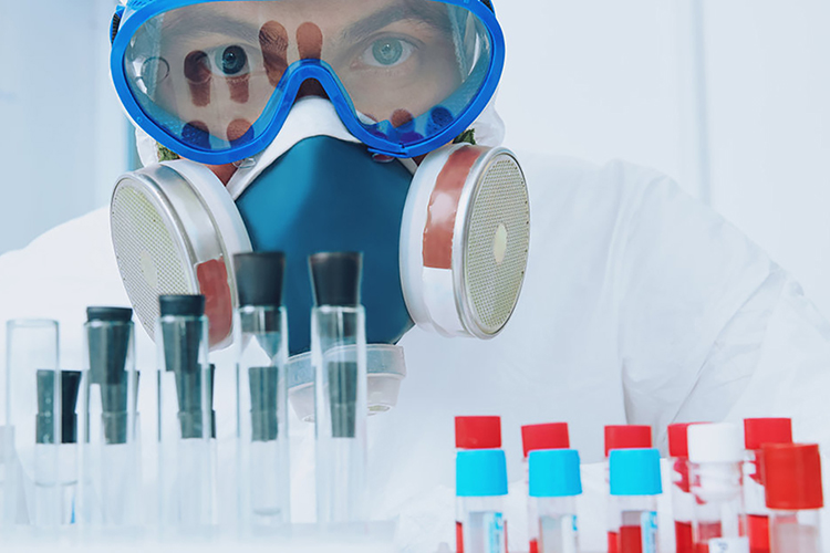 Scientist in respirator, goggles and other protective gear looking at camera over a lab bench of sample tubes - idea of HPAPIs