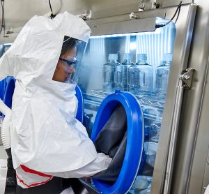 woman in white hazard suit and respiratory gear with hands in a vacuum chamber