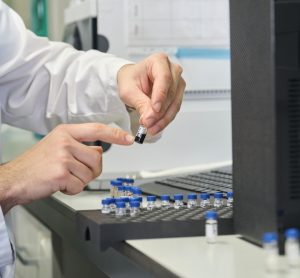 Scientist in a white lab coat putting vial with sample into autosampler of HPLC system.