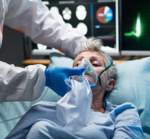 Doctor in full PPE holding an oxygen mask over the nose and mouth of an elderly woman with COVID-19 in a hospital bed