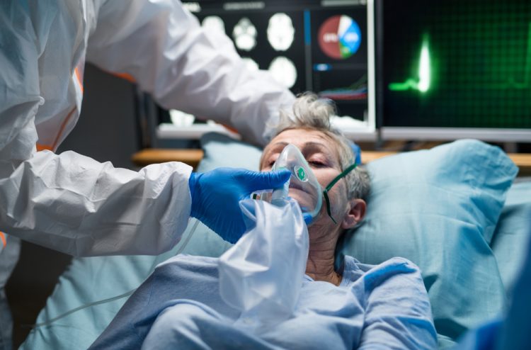 Doctor in full PPE holding an oxygen mask over the nose and mouth of an elderly woman with COVID-19 in a hospital bed