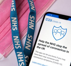 Phone screen displaying the introduction page to the NHS COVID-19 app, next to a lanyard labelled with NHS and a pink facemask