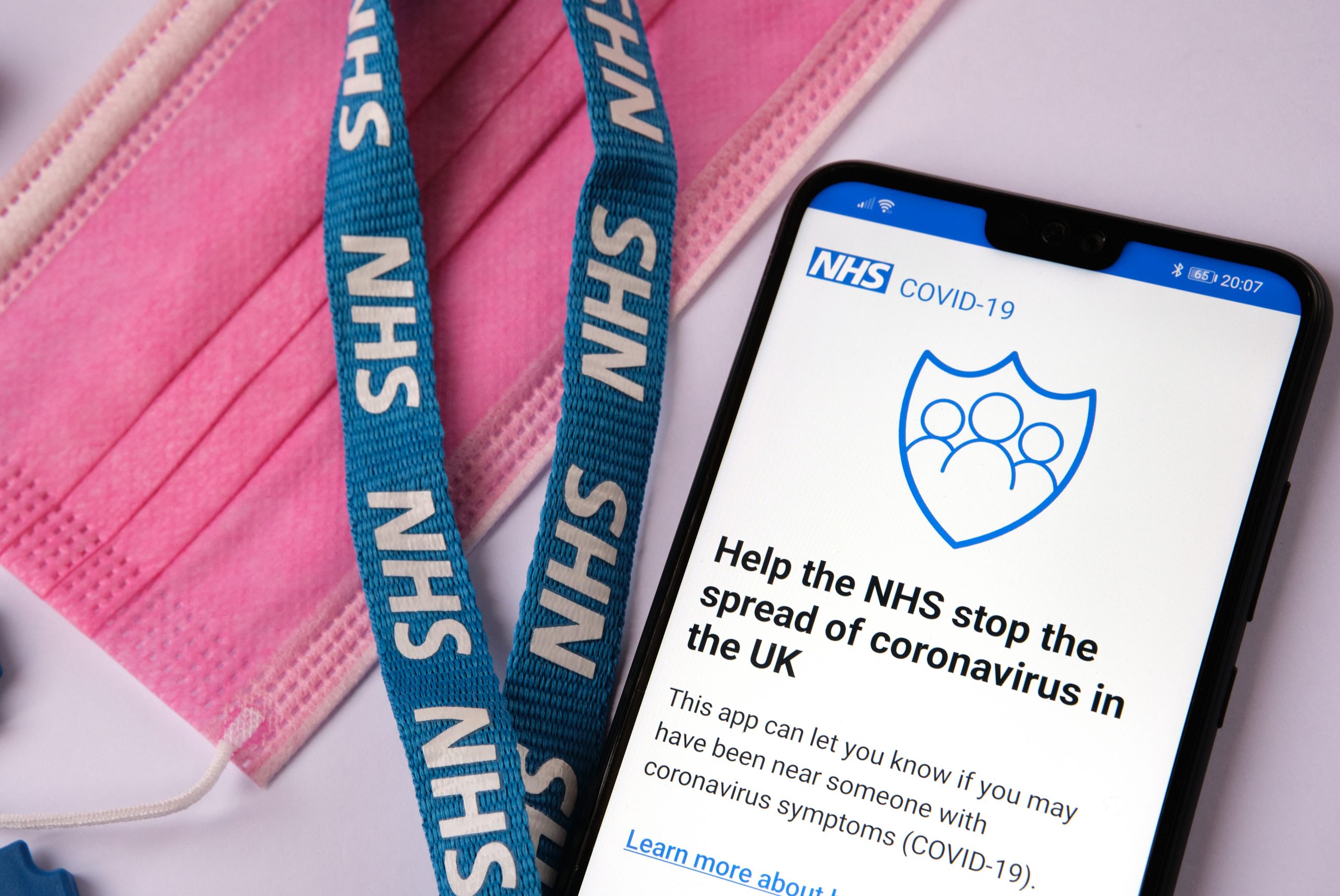 Phone screen displaying the introduction page to the NHS COVID-19 app, next to a lanyard labelled with NHS and a pink facemask
