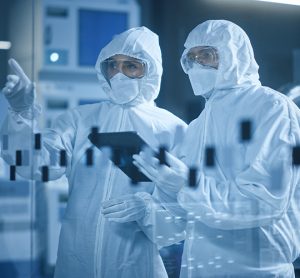 Environmental monitoring/microbiology/aseptic processing concept - factory cleanroom: engineer and scientist wearing coveralls, talking and using tablet computer