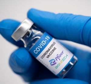 Gloved hand holding a vial labelled 'COVID-19 Vaccine' with Pfizer and BioNTech logos [Credit: malazzama / Shutterstock.com].
