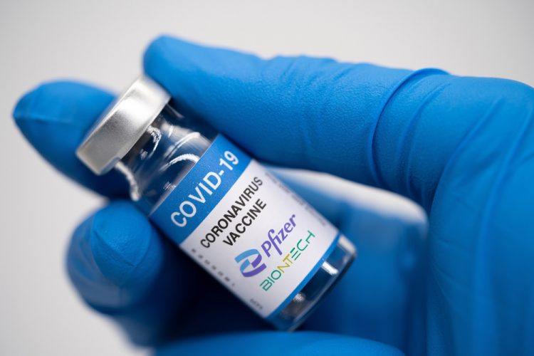 Gloved hand holding a vial labelled 'COVID-19 Vaccine' with Pfizer and BioNTech logos [Credit: malazzama / Shutterstock.com].