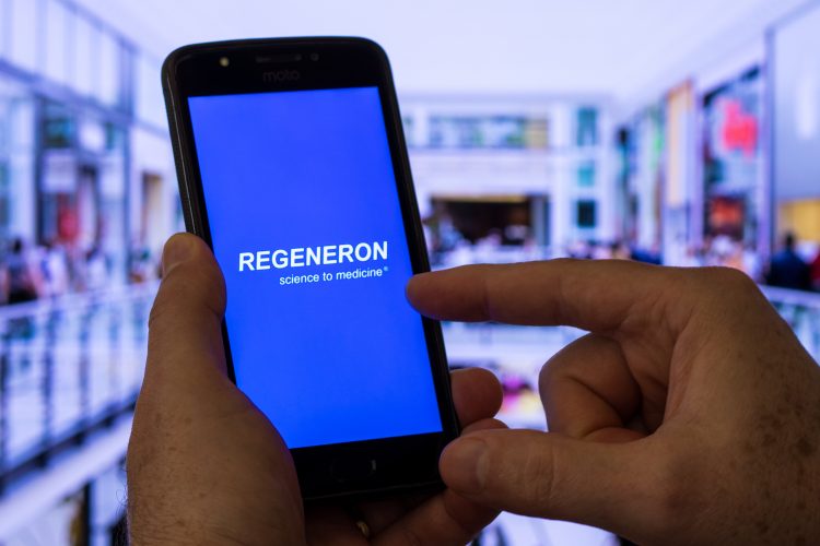 Phone with regeneron logo on it in front of blurred laboratory background [Credit: adrianosiker.com/Shutterstock.com].