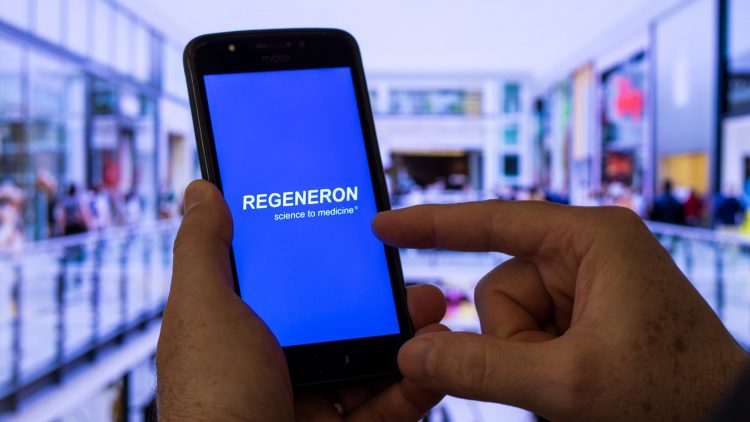 Phone with regeneron logo on it in front of blurred laboratory background [Credit: adrianosiker.com/Shutterstock.com].