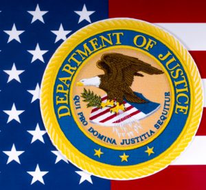 US Department of Justice logo on top of an American Flag [Credit: US-Department-of-Justice].
