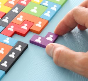 close up of person's hand sliding a wooden puzzle block with people icons on it into a space in a larger puzzle - idea of appointments or recruitment