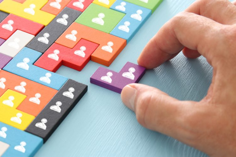 close up of person's hand sliding a wooden puzzle block with people icons on it into a space in a larger puzzle - idea of appointments or recruitment