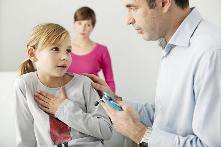 female child with hand over chest - indicating trouble breathing - next to doctor in white room