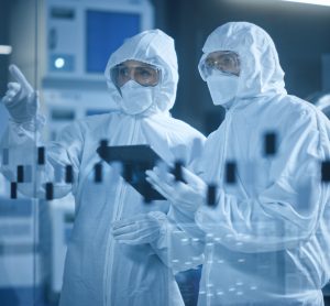 Two people wearing cleanroom garments and gloves looking at a computer tablet in a manufacturing cleanroom facility