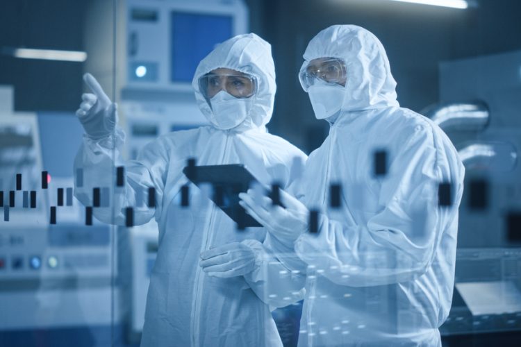 Two people wearing cleanroom garments and gloves looking at a computer tablet in a manufacturing cleanroom facility