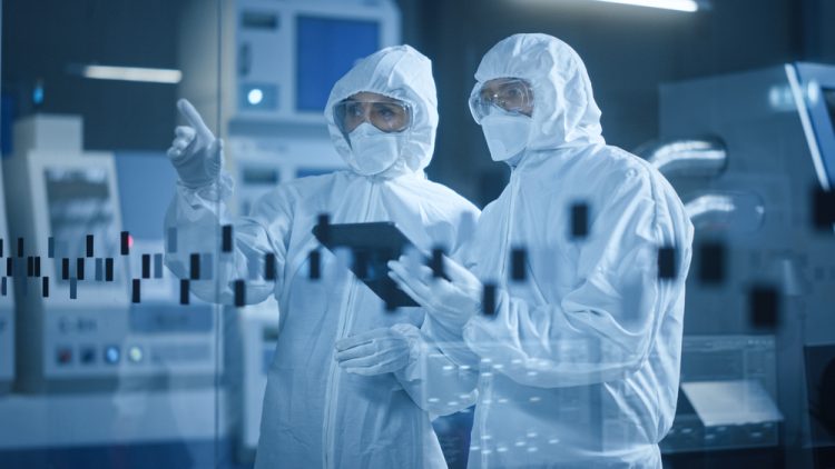 Two people wearing cleanroom garments and gloves looking at a computer tablet in a manufacturing cleanroom facility